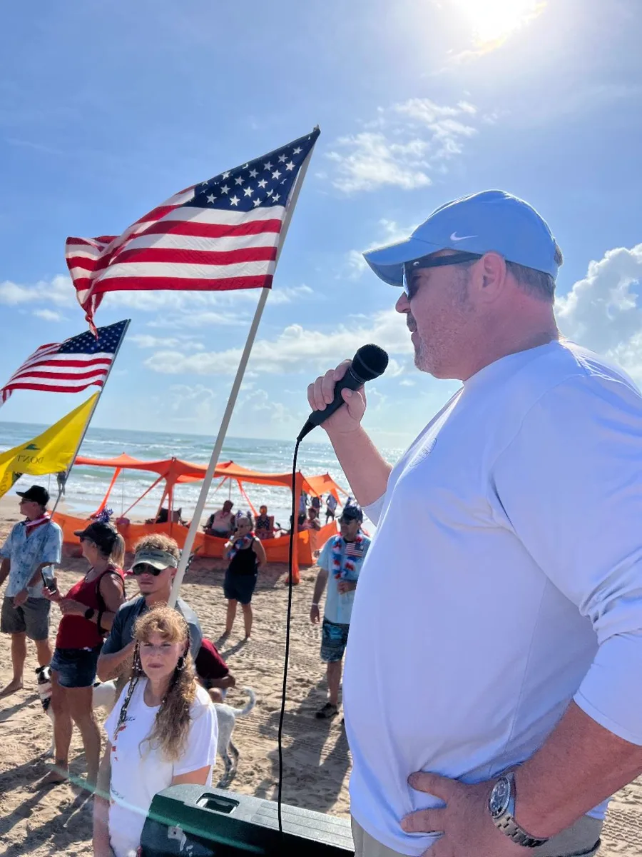 Patrick McNulty speaking into a microphone on the beach at South Padre Island, with American flags and the Gulf behind him.