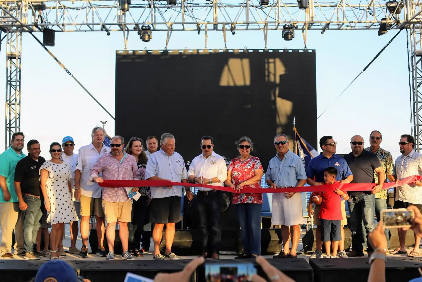 A ribbon cutting on stage at South Padre Island, with residents and officials holding the ribbon and a crowd photographing it.